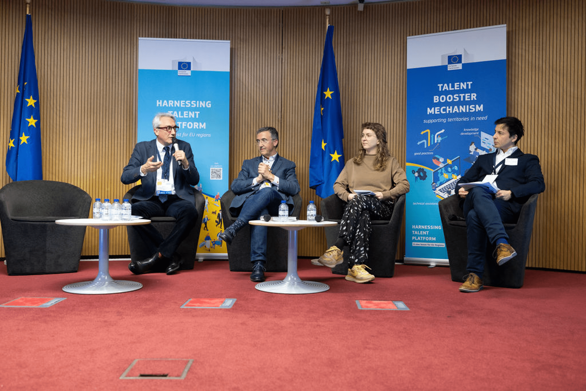 Four people sit on a panel stage with two EU flags and banners.